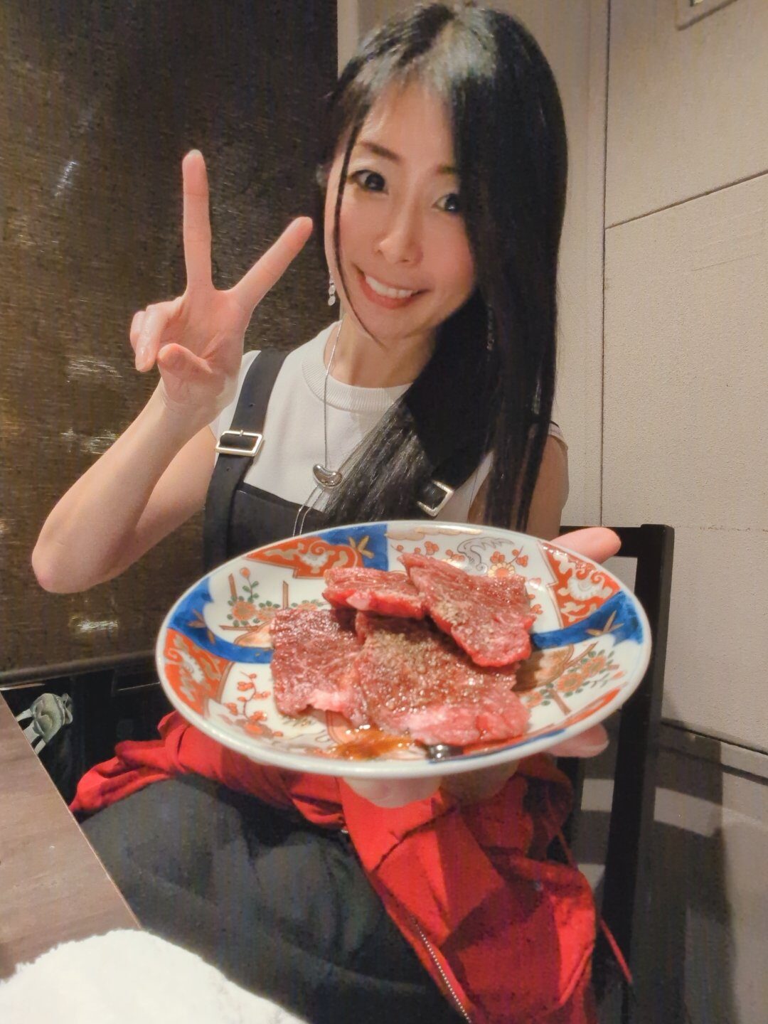 笑顔の長谷川香枝が右手でピースして、左手で生肉の小皿をこちらに見せている