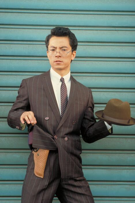 熱中時代・刑事編の水谷豊、左手にハットを持って、右手でジャケットの裾を持ち上げている